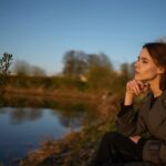 Person observing nature beside a lake while reflecting on creative ideas