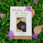Writing Down the Bones by Natalie Goldberg surrounded by purple flowers, fallen leaves, and green grass.